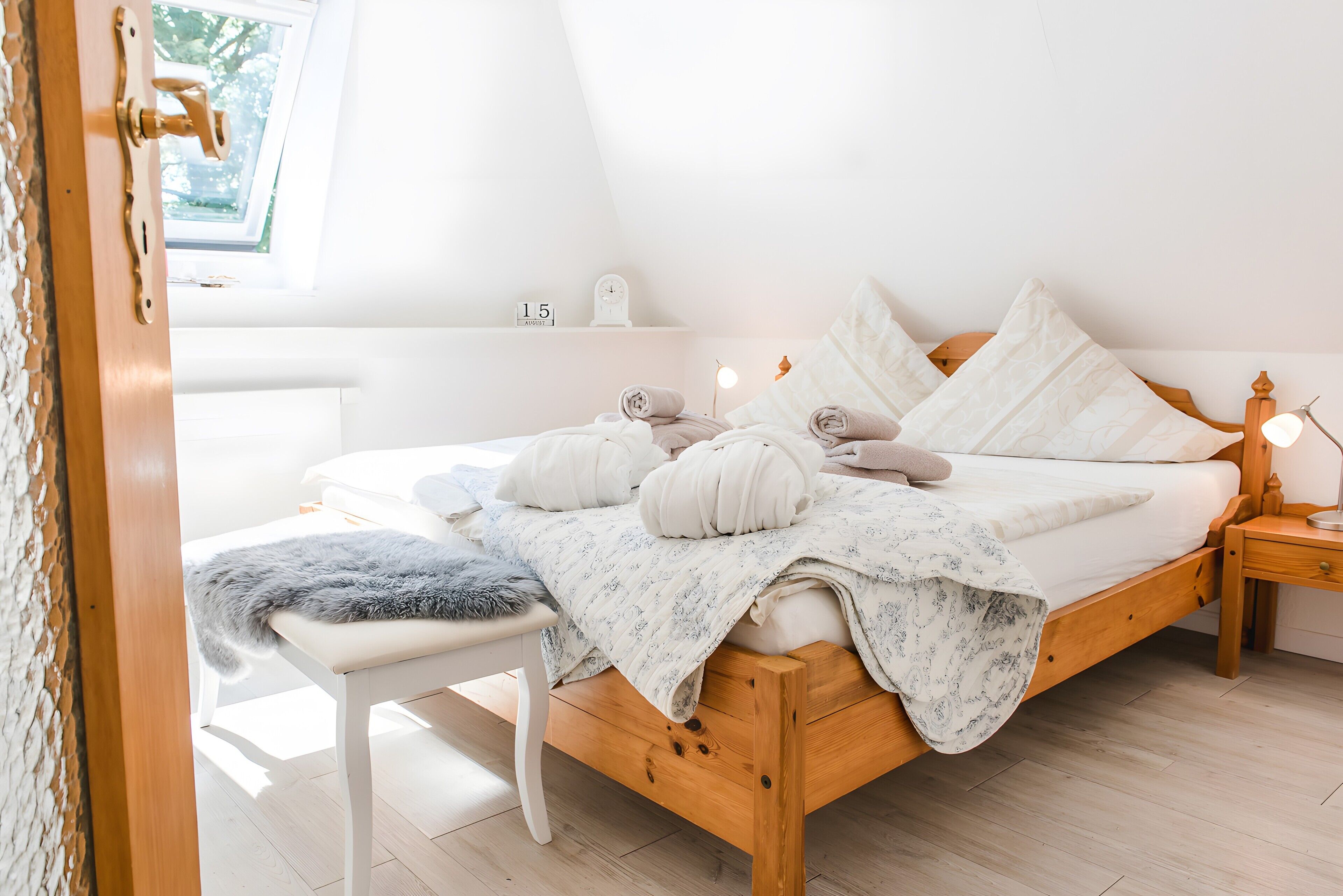 Une chambre à coucher avec un lit en bois, du linge de lit blanc, une table de chevet, une petite étagère et une fenêtre.