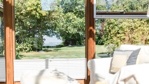 A room with a view of a grassy area and trees, a white chair, and a wooden table. - Holiday Home 'Am Meer' with Sea View, Private Terrace and Wi-Fi (Langballig)