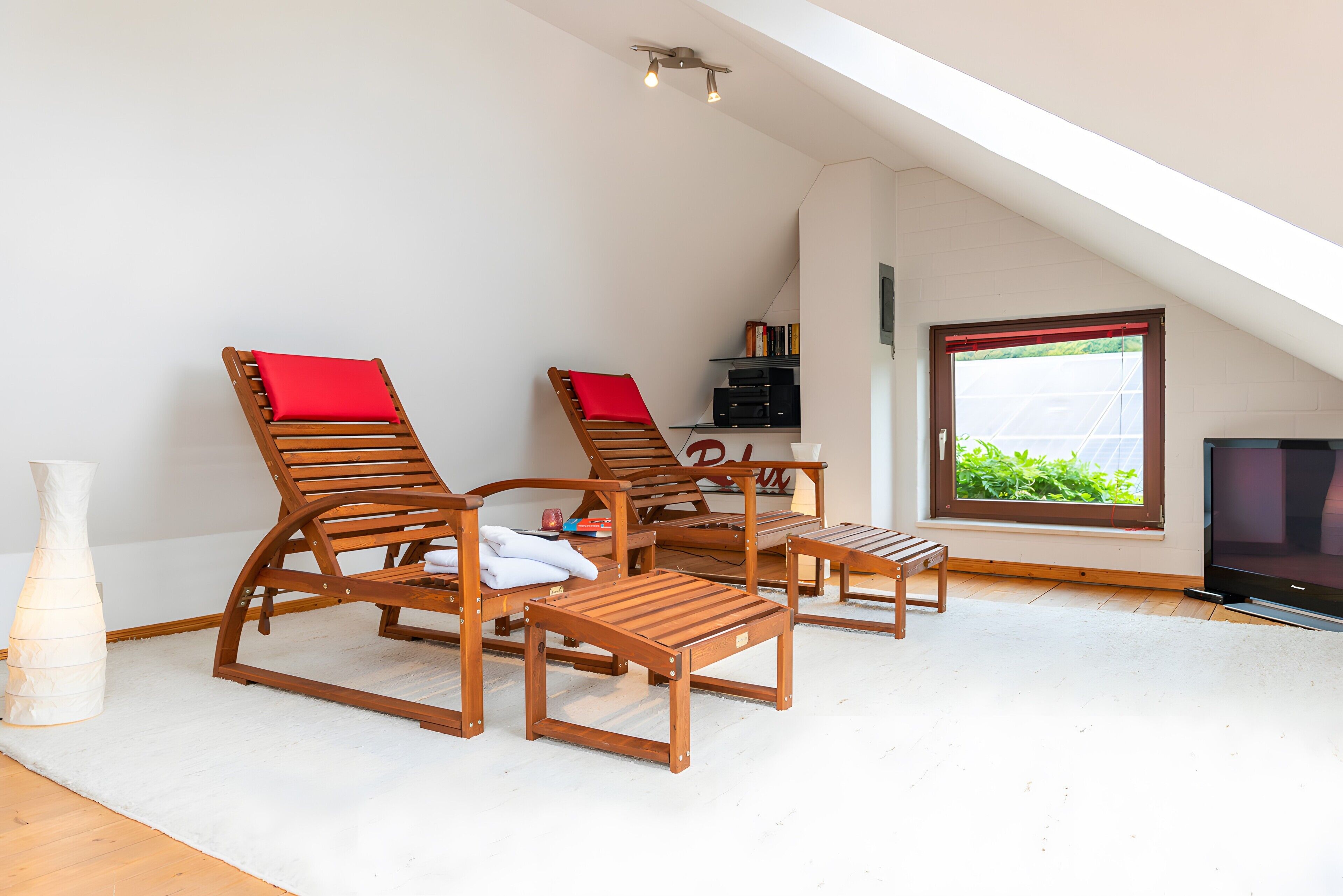 A room with a sloped ceiling, wooden lounge chairs with red cushions, a small table, a bookshelf, and a television.