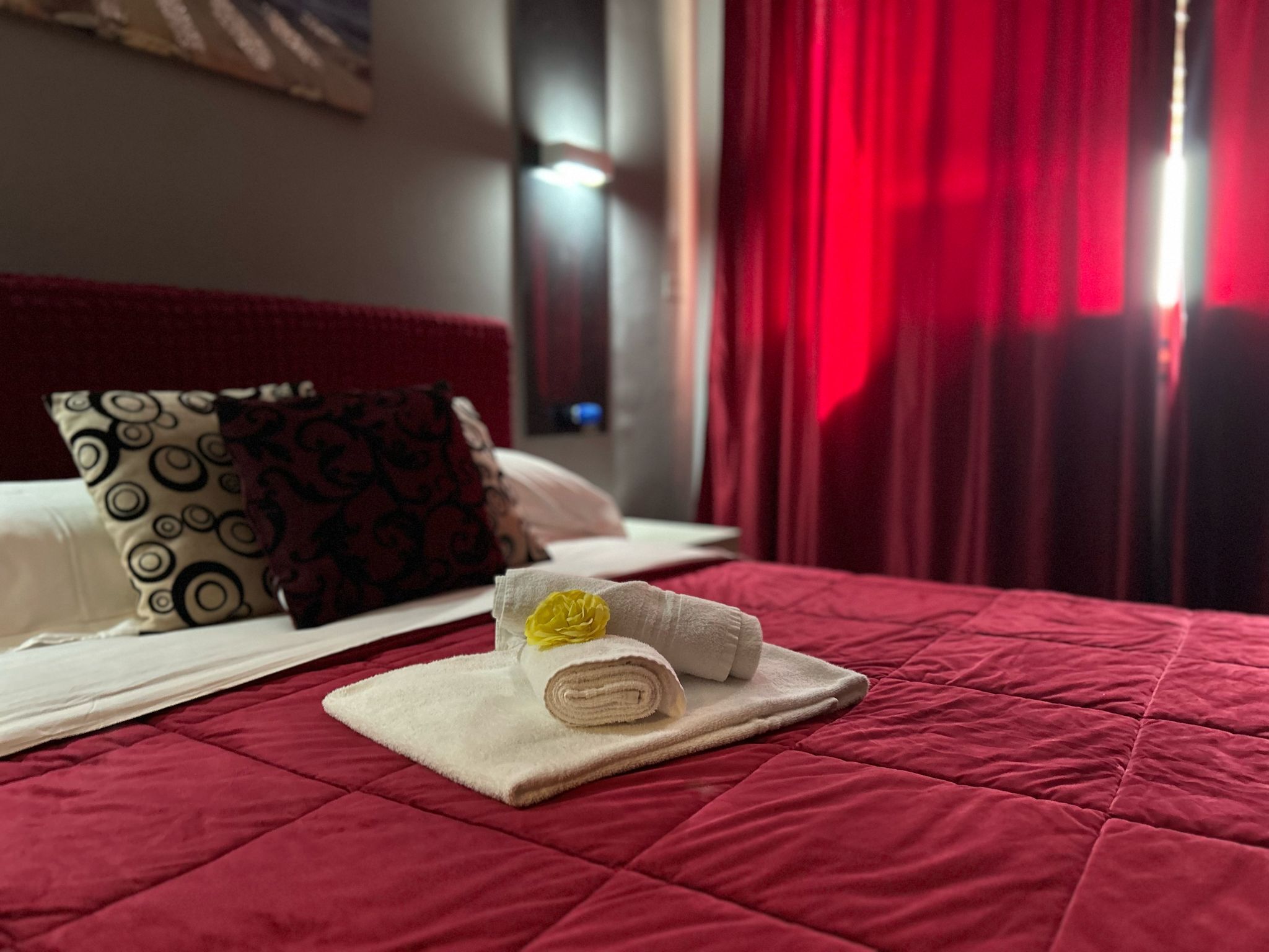 Cama bem arrumada com colcha vermelha, travesseiro branco com padrão circular e toalha branca enrolada com flor amarela.