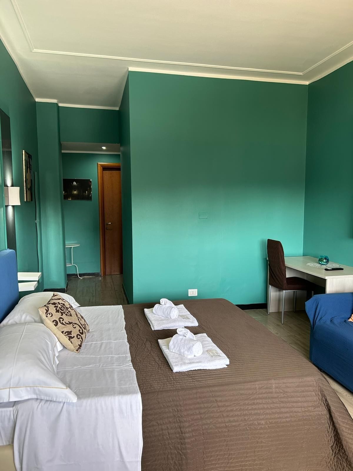 Una camera d'albergo con pareti color acquamarina, un letto con biancheria bianca e marrone, una scrivania con una sedia e un piccolo tavolo con una lampada.