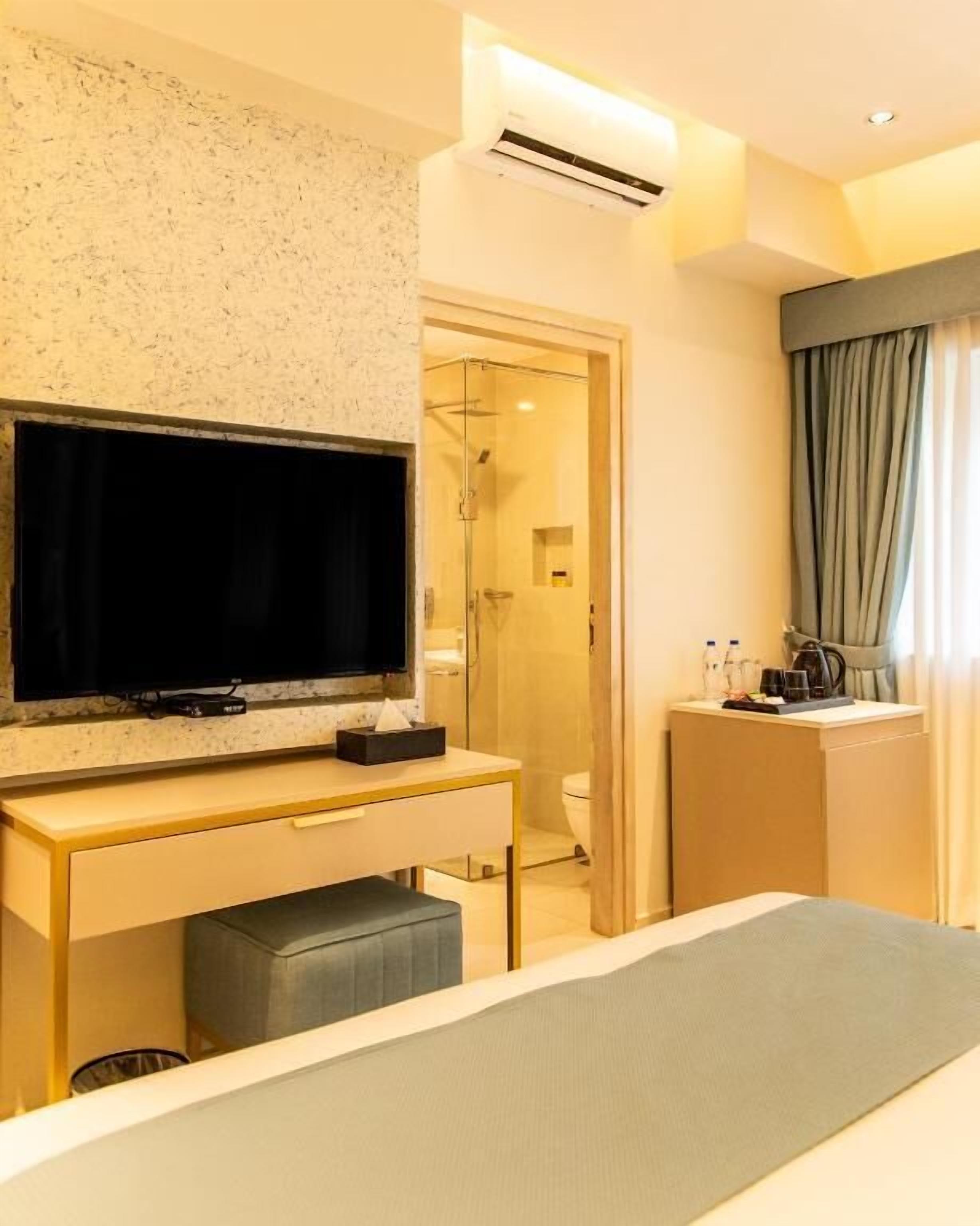 A hotel room with a bed, a TV on a wooden desk, a shower area, and a window with curtains.