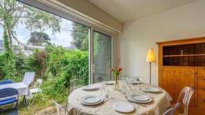 A round table with a beige tablecloth, surrounded by clear chairs, set for dining with plates, cutlery, and a vase with red flowers.