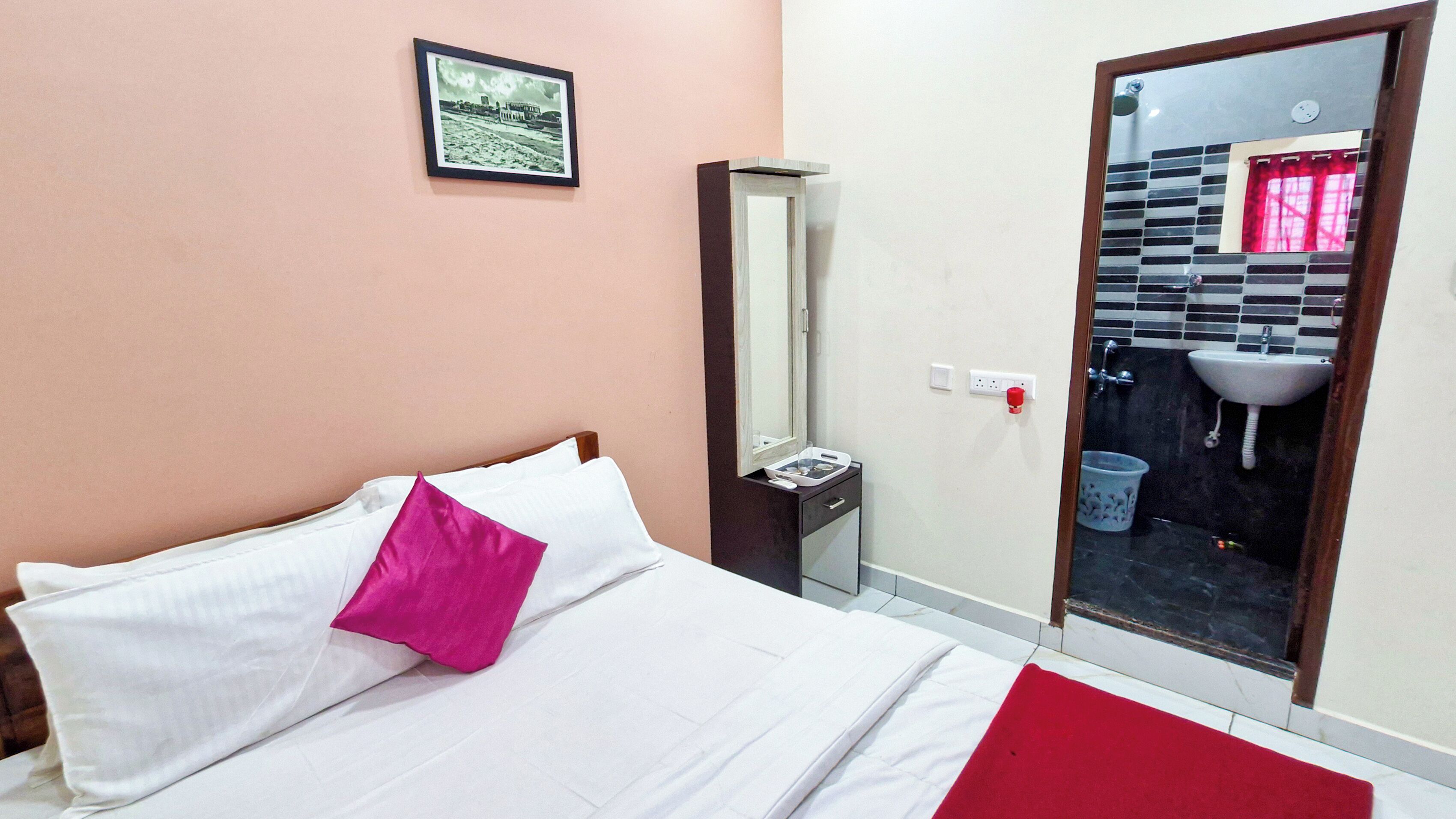 A hotel room with a bed, a bedside table, a mirror, a red throw pillow, a red blanket, and a view of a bathroom with a sink and a tiled wall.