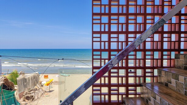 A beach view from a balcony with a red lattice structure, a wooden bench, and a clear blue sky.