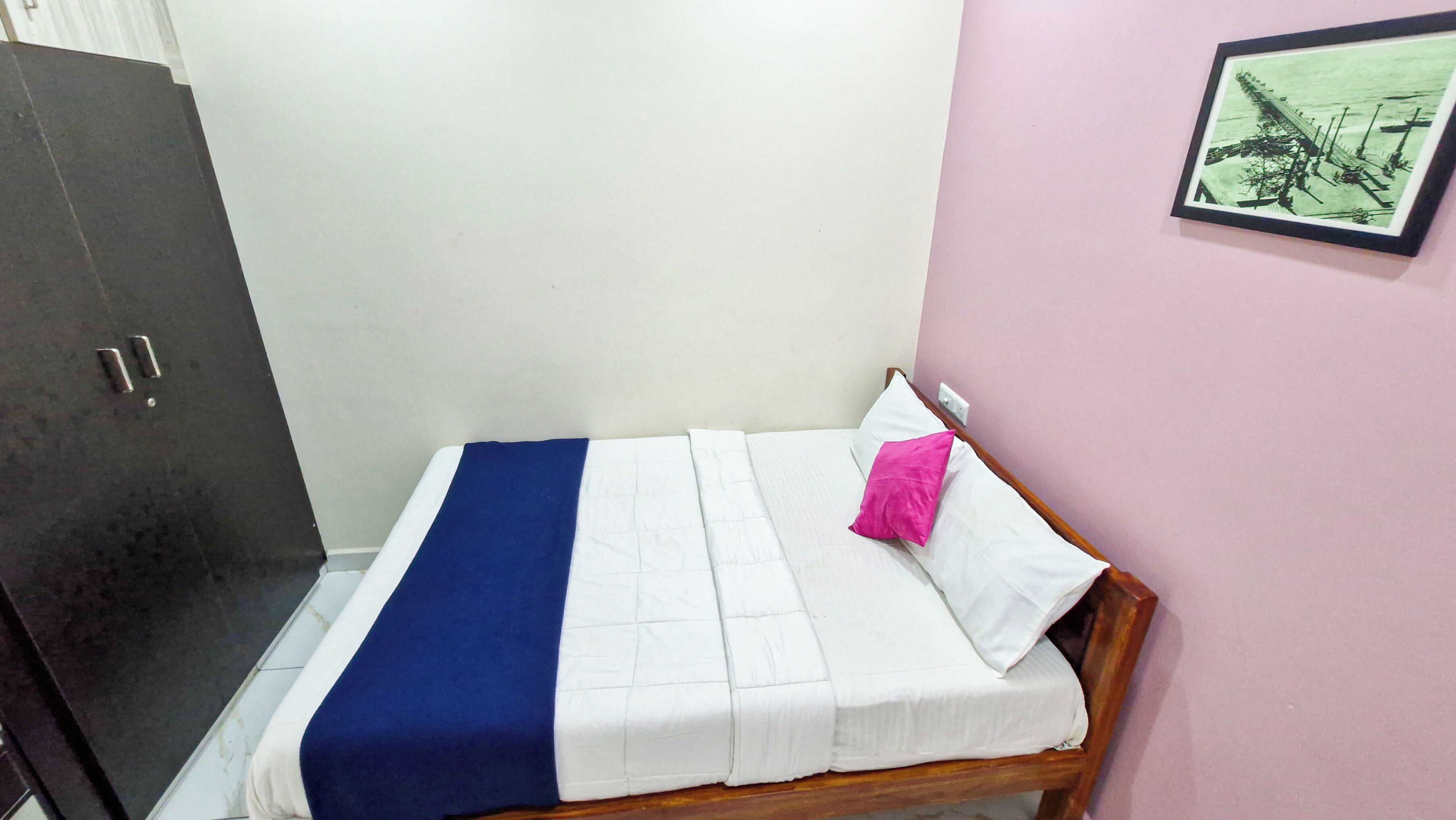 A small, neatly made bed with a blue and white bedspread and a pink pillow, a wooden wardrobe, and a framed picture on the wall.