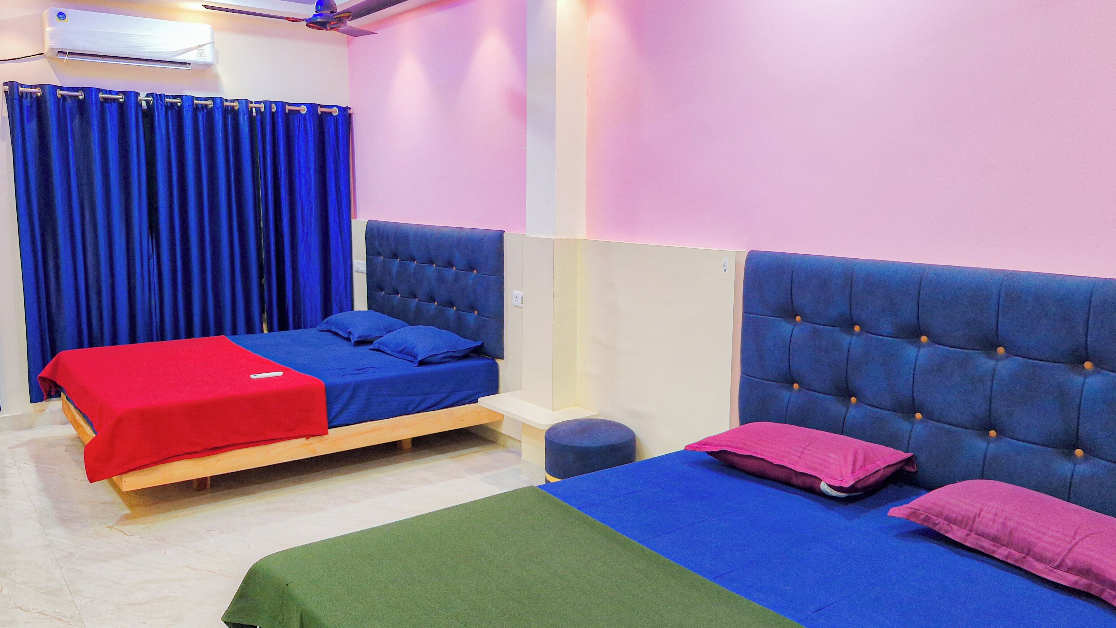 A room with two beds, each with a blue headboard and pillows, a red blanket on one bed, and a green blanket on the other. There is a small round table and a blue curtain.