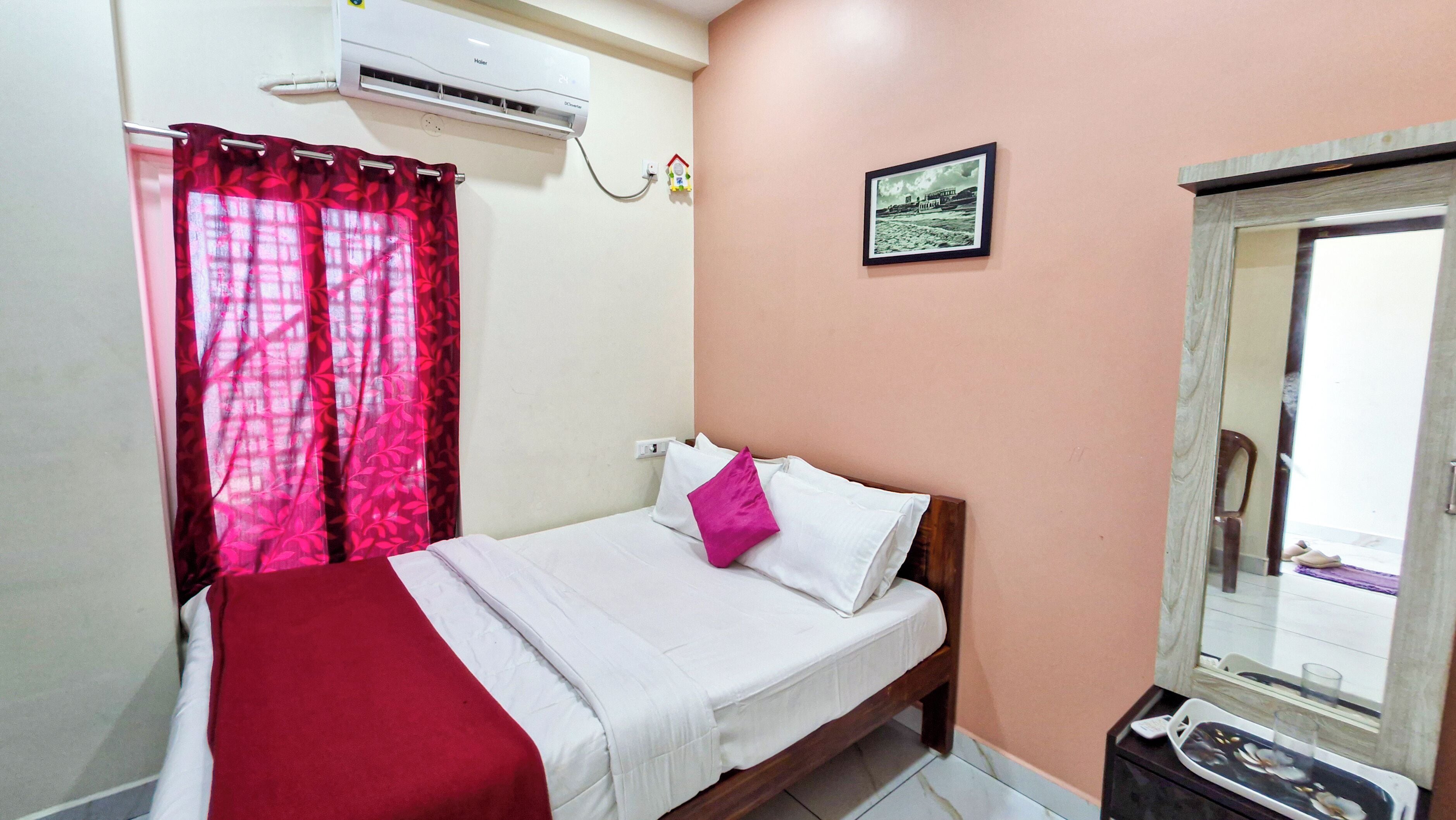 Quarto com cama, colcha vermelha e branca, travesseiro rosa, ar-condicionado fixado na parede, quadro emoldurado e janela com vista.