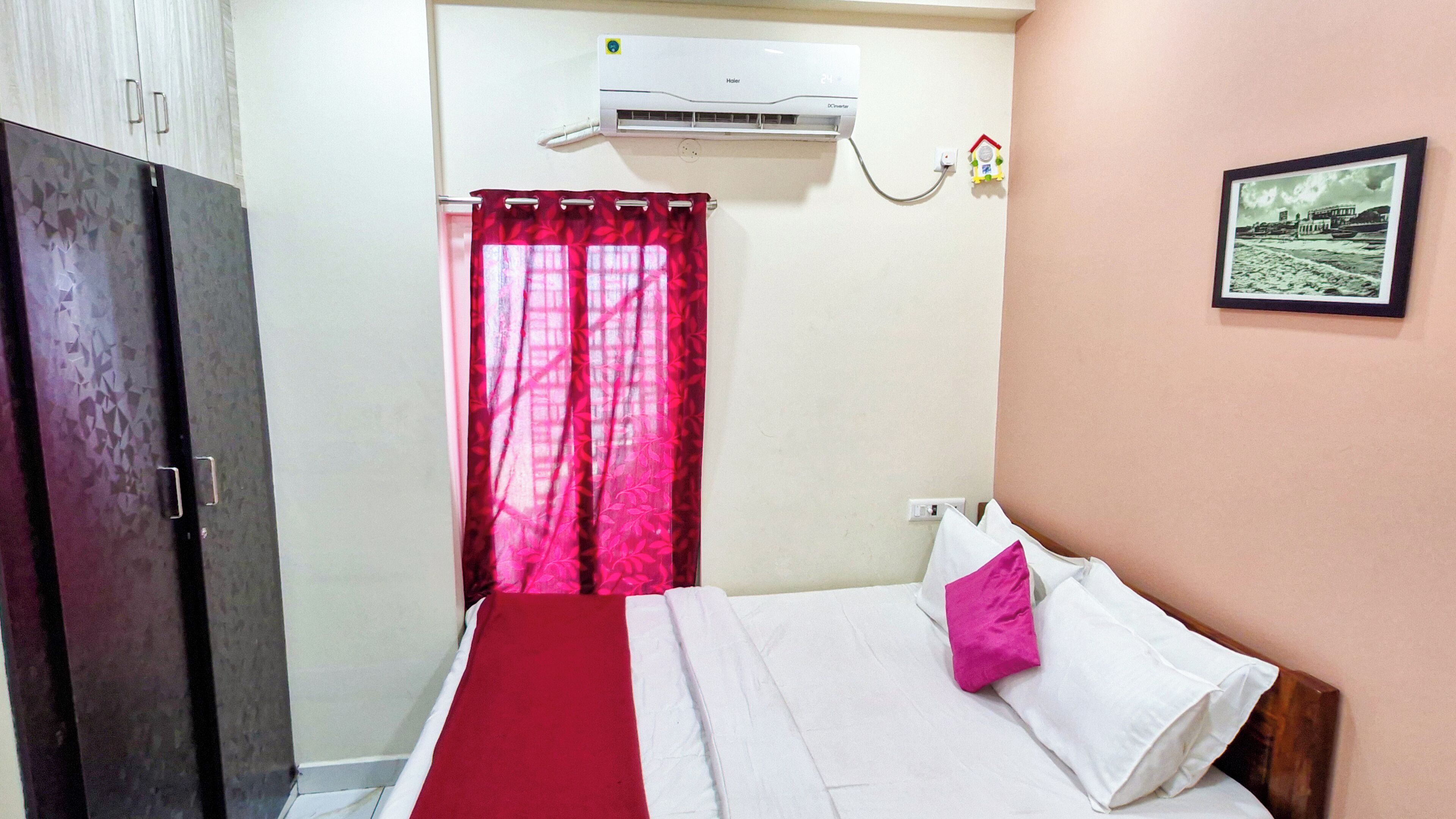 Un dormitorio con cama, armario, ventana con cortinas rojas, aire acondicionado y un cuadro en la pared.