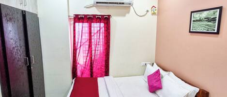 Un dormitorio con cama, armario, ventana con cortinas rojas, aire acondicionado y un cuadro en la pared.