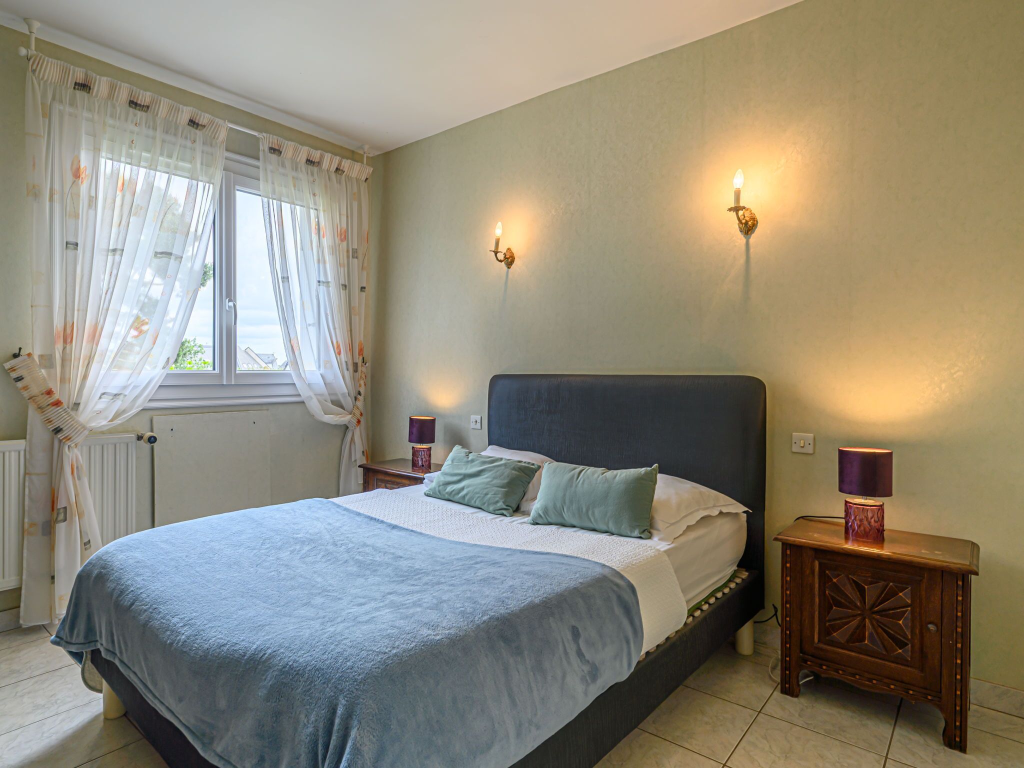 Une chambre à coucher avec un lit, deux lampes de chevet, une fenêtre avec des rideaux et un radiateur.