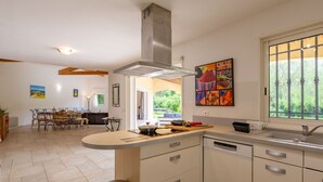 A modern kitchen with a central island, a dining area with a wooden table and chairs, and a view of greenery through a window. - La Pimaleanne by Interhome (Roquefort les Pins)