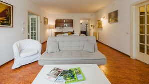 A living room with a grey sofa, a white armchair, and a coffee table with magazines. There is a wooden headboard with a 'YES' sign above a bed in the background. - Lido Versilia by Interhome (Lido di Camaiore)