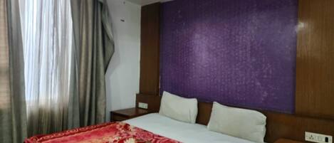 A hotel room with a bed, a nightstand, a purple accent wall, and a window with curtains.