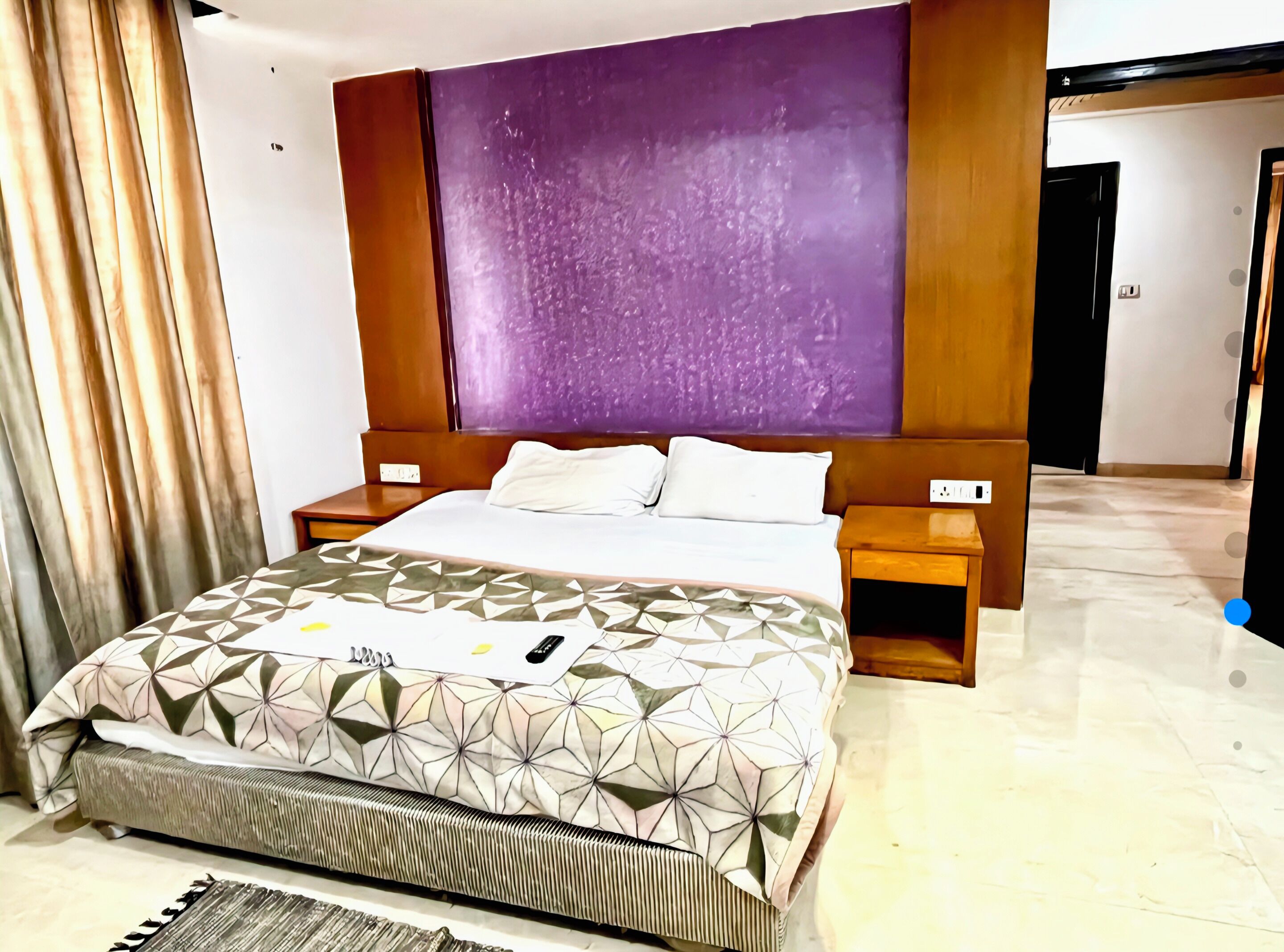 A hotel room with a large bed, a headboard with a purple panel, two bedside tables, and a window with curtains.