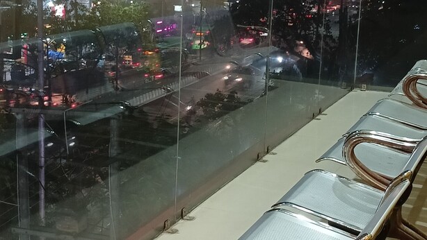 A long, narrow seating area with metal and wooden chairs, a glass partition, and a view of a cityscape at night. - Sacred Stay (Guwahati)