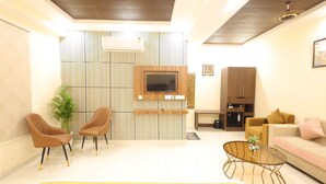 A modern living room with a flat-screen TV mounted on a wooden panel wall, two leather armchairs, a glass coffee table, a sofa, and a kitchen area with a refrigerator and cabinets. - GP Palace (Lucknow)