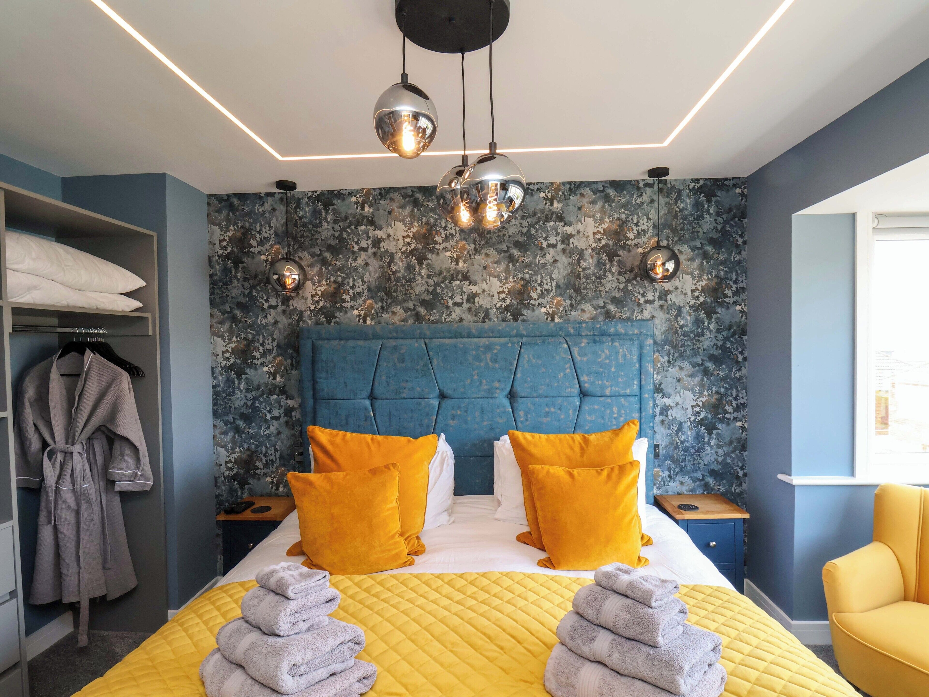 A modern bedroom with a blue headboard, two yellow pillows, a bed with a yellow quilt, and a stack of towels.
