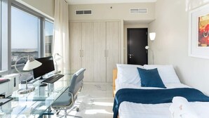 Una habitación de hotel moderna con un escritorio de vidrio, una cama con ropa de cama azul, un ventanal grande y una puerta que da a un armario.