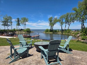 Patio con sillas Adirondack y un brasero con vista a un lago, muelle y barcos.