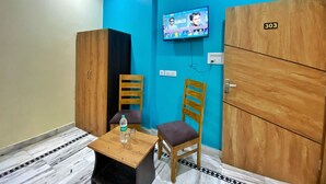 Een kamer met een blauwe wand, een houten garderobe, een aan de muur bevestigde televisie, een kleine tafel met een fles en glazen, en een stoel.
