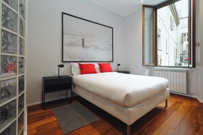 A bedroom with a large bed, a nightstand, a bedside table, a framed picture on the wall, a window with a view of buildings, and a storage unit with clear plastic doors.