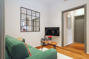 Un salon avec un canapé vert, une petite table en bois sur laquelle est posé un vase, un téléviseur fixé au mur et une porte donnant sur une autre pièce.