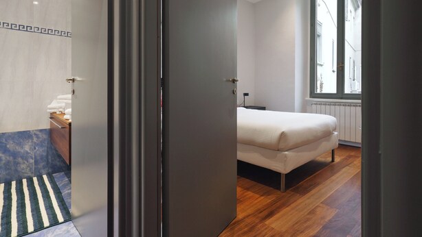A modern bedroom with a large bed, a wooden floor, a radiator, and a window with a view of buildings. - Stylish 2BR flat in the city centre (Milan)
