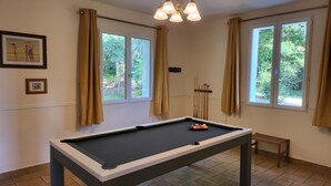 A pool table with a billiard cue rack and balls, two windows with curtains, and framed pictures on the wall.