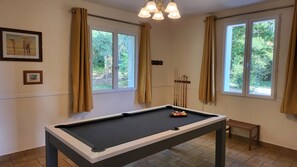 Uma mesa de bilhar com suporte para tacos e bolas, duas janelas com cortinas, e quadros nas paredes.