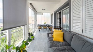 Een balkon met een grijze bank, gele kussens, een eettafel met stoelen, en potplanten.