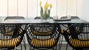 Een eethoek met een tafel voor vier personen, omringd door zwarte rieten stoelen met gele kussens. Op de tafel staat een glazen vaas met gele tulpen.