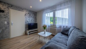 Una sala de estar moderna con un sofá gris, una mesa de centro blanca, un televisor de pantalla plana y una ventana con vistas a zonas verdes.