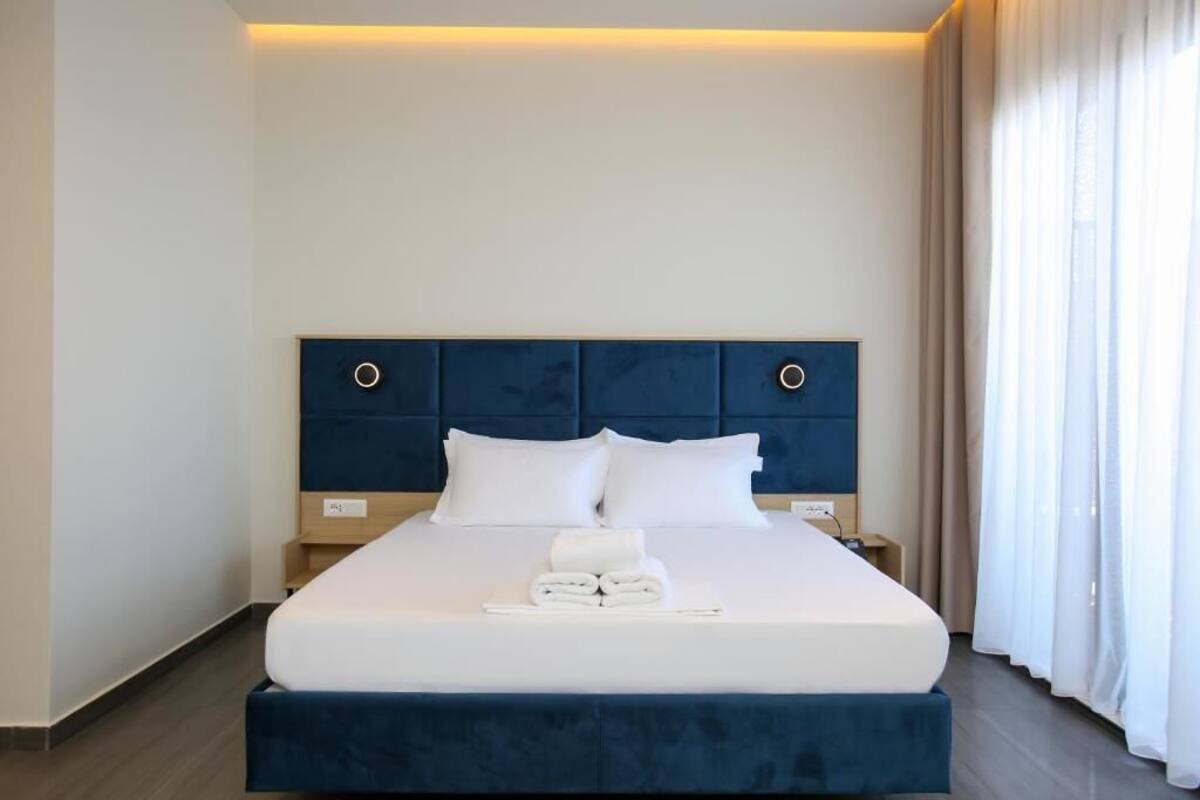 A neatly made bed with white linens and a blue headboard, a folded towel on the bed, and a bedside table with a lamp.