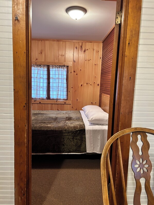 A small, wooden-paneled room with a single bed, a window with plaid curtains, and a ceiling light.