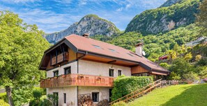 A two-story chalet with a wooden balcony, a stone facade, and a mountain backdrop.
