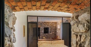 Une pièce avec un mur en pierre, un lit avec une literie à motifs, une petite table et une lampe.