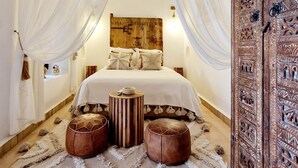 A bedroom with a bed, two stools, a decorative wooden headboard, and a large wooden door with intricate carvings. - Riad Dar El Mahja (Marrakech)