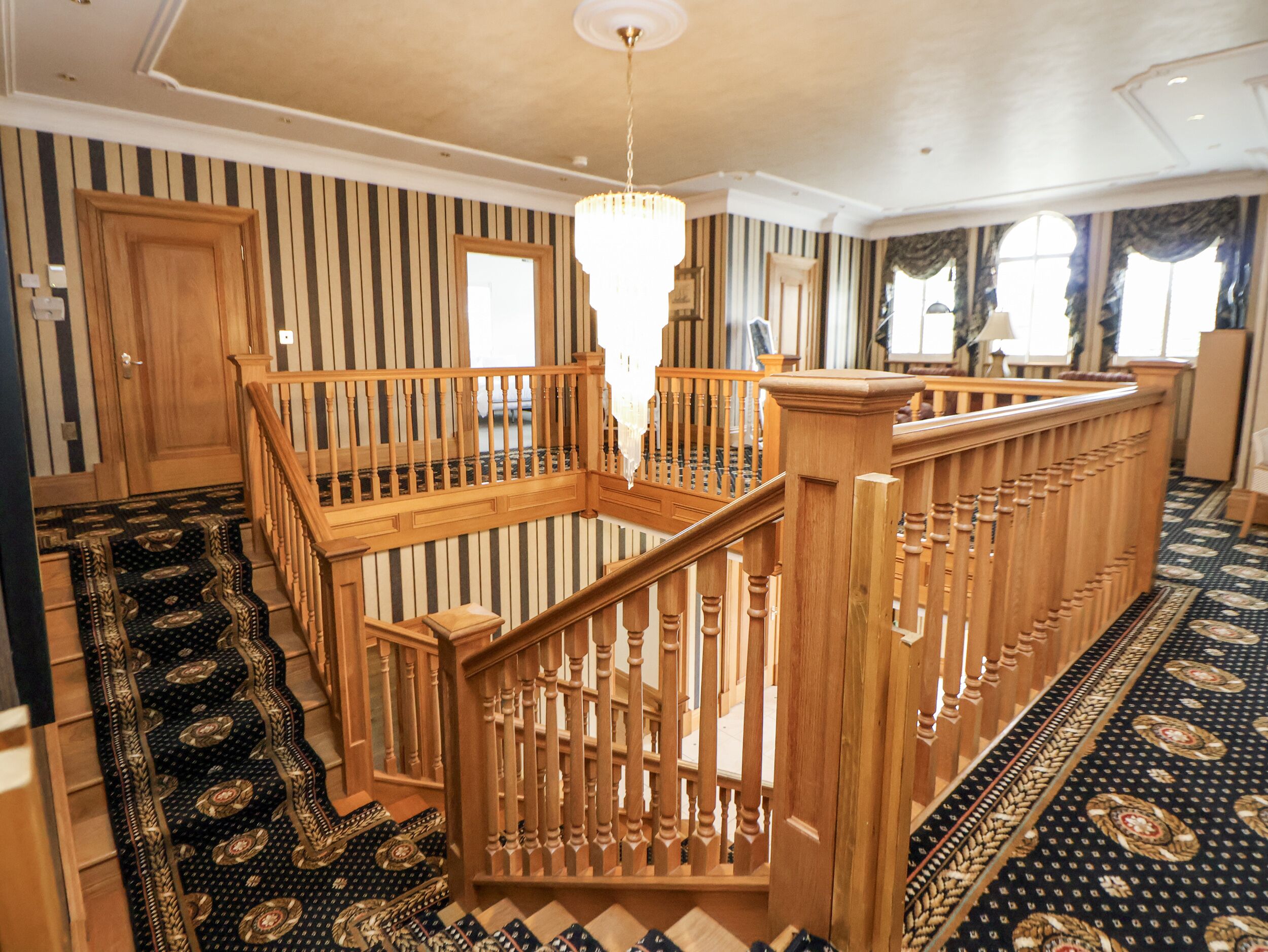 Una habitación amplia con escalera de madera, candelabro y alfombra estampada.