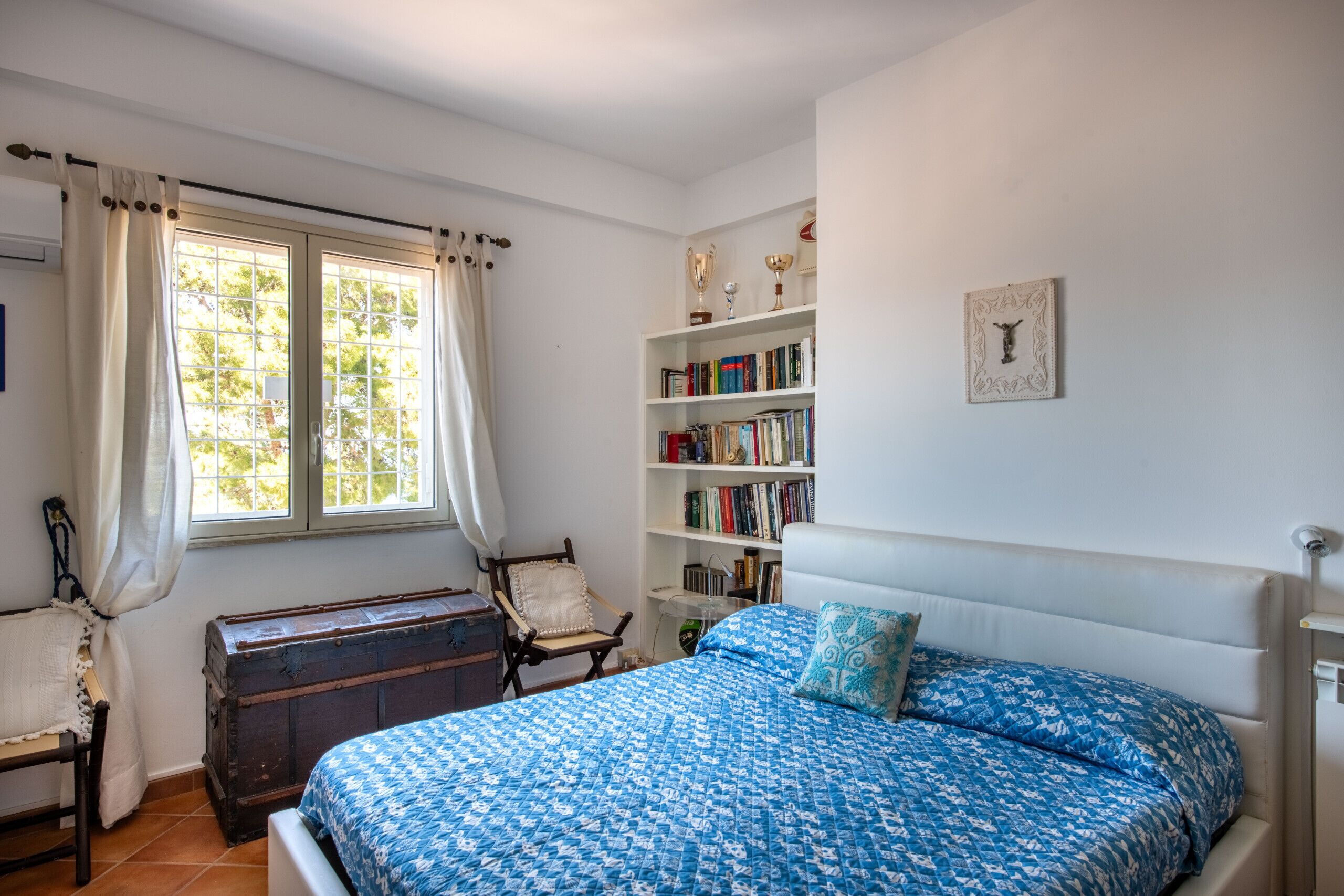 A bedroom with a bed, a bookshelf, a window with curtains, and a wooden chest.