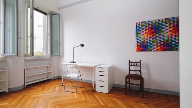 Une pièce avec un plancher en bois, un bureau blanc, une chaise et un tableau abstrait aux couleurs vives accroché au mur.