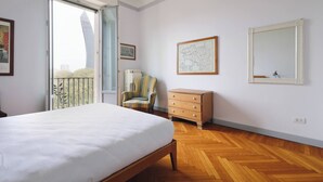 Une chambre à coucher comprenant un lit, une commode, une chaise et une grande fenêtre donnant sur un immeuble.