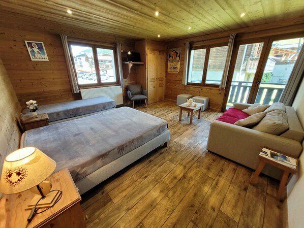 A cozy room with a bed, a sofa, a small table, and a chair. There are posters on the walls and a window with a view.