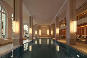 Une grande piscine intérieure dotée d’un espace de détente central, entourée de chaises longues et surmontée d’un haut plafond avec des luminaires encastrés.