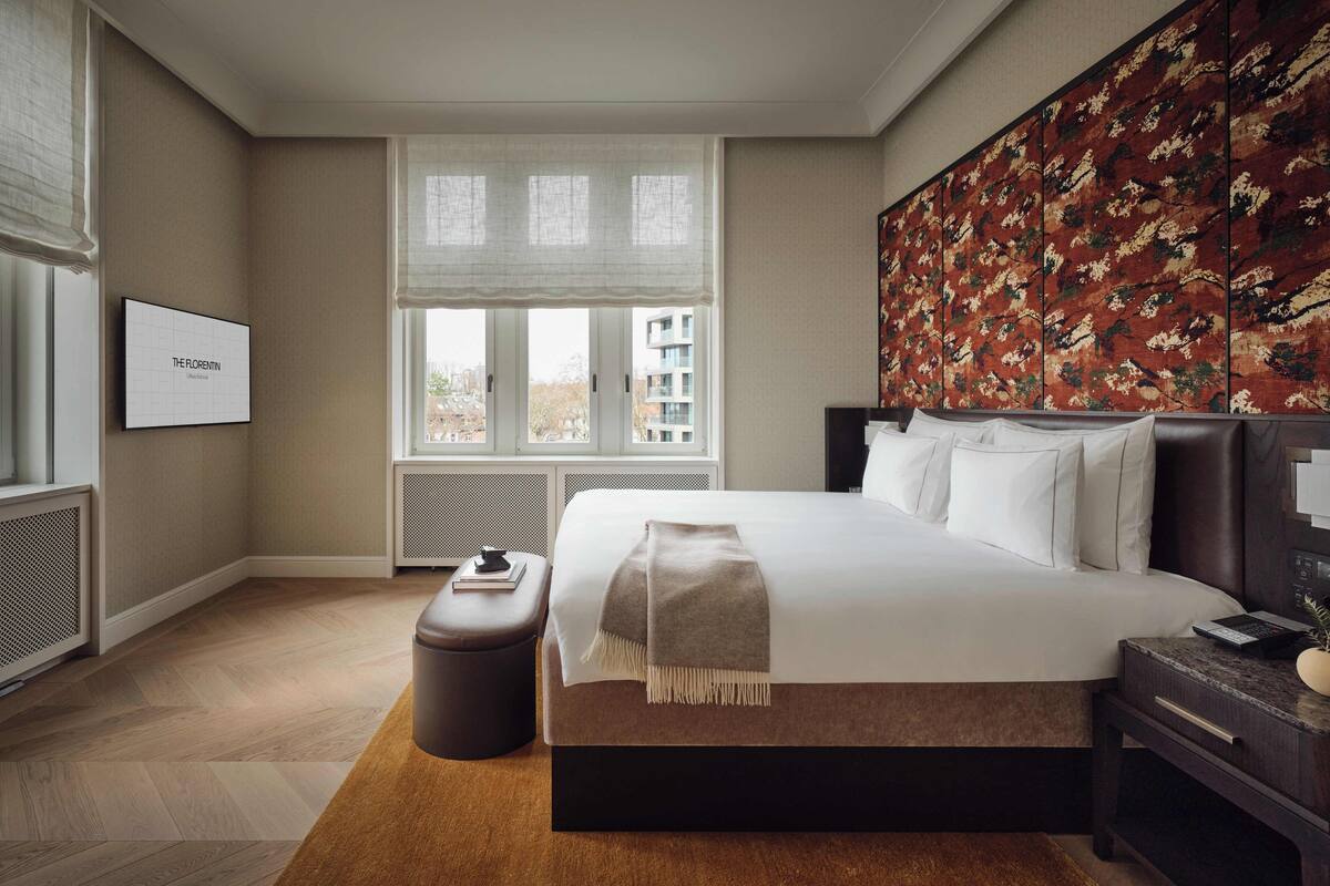 A hotel room with a large bed, a nightstand, a window with blinds, a radiator, and a wall-mounted artwork.