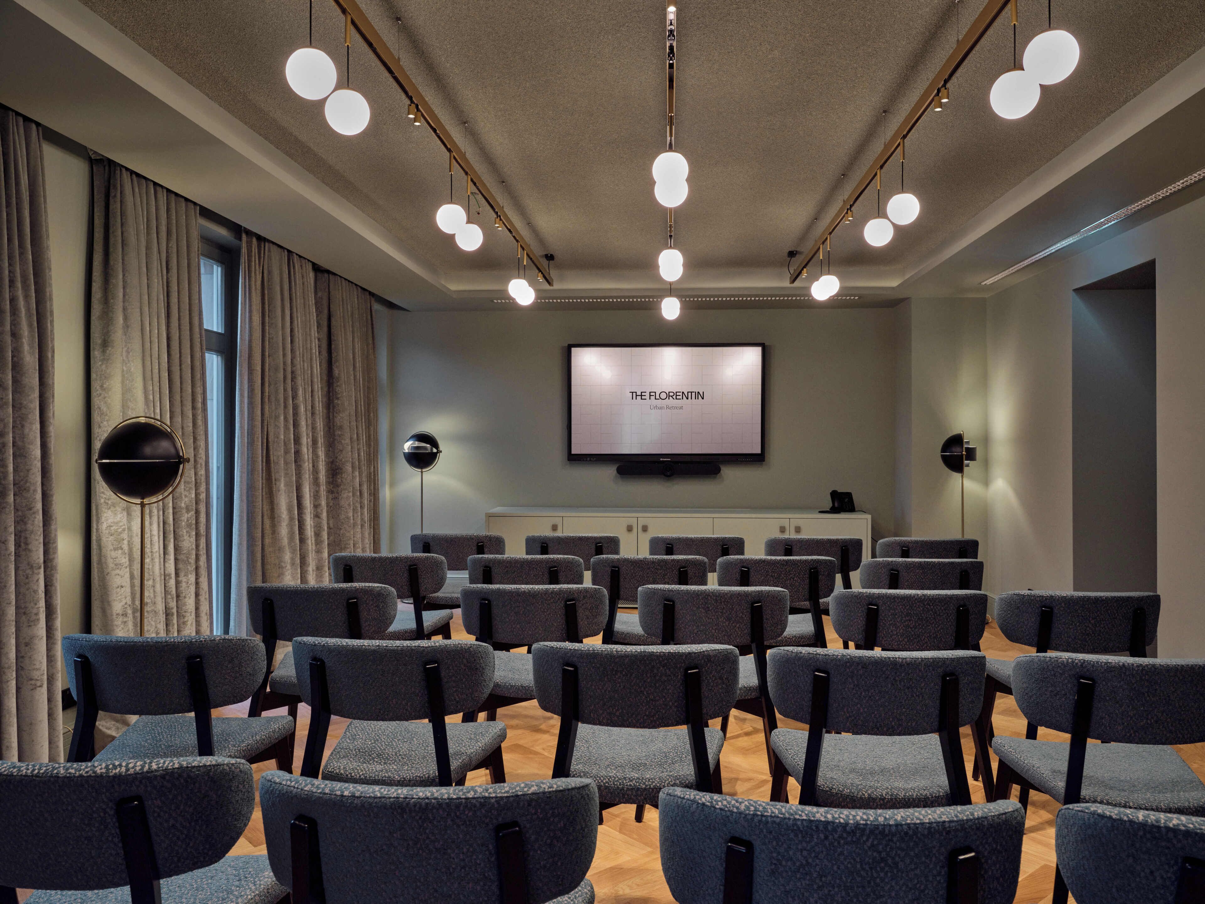 Une salle de conférence avec des rangées de chaises, un grand écran et des luminaires au plafond.