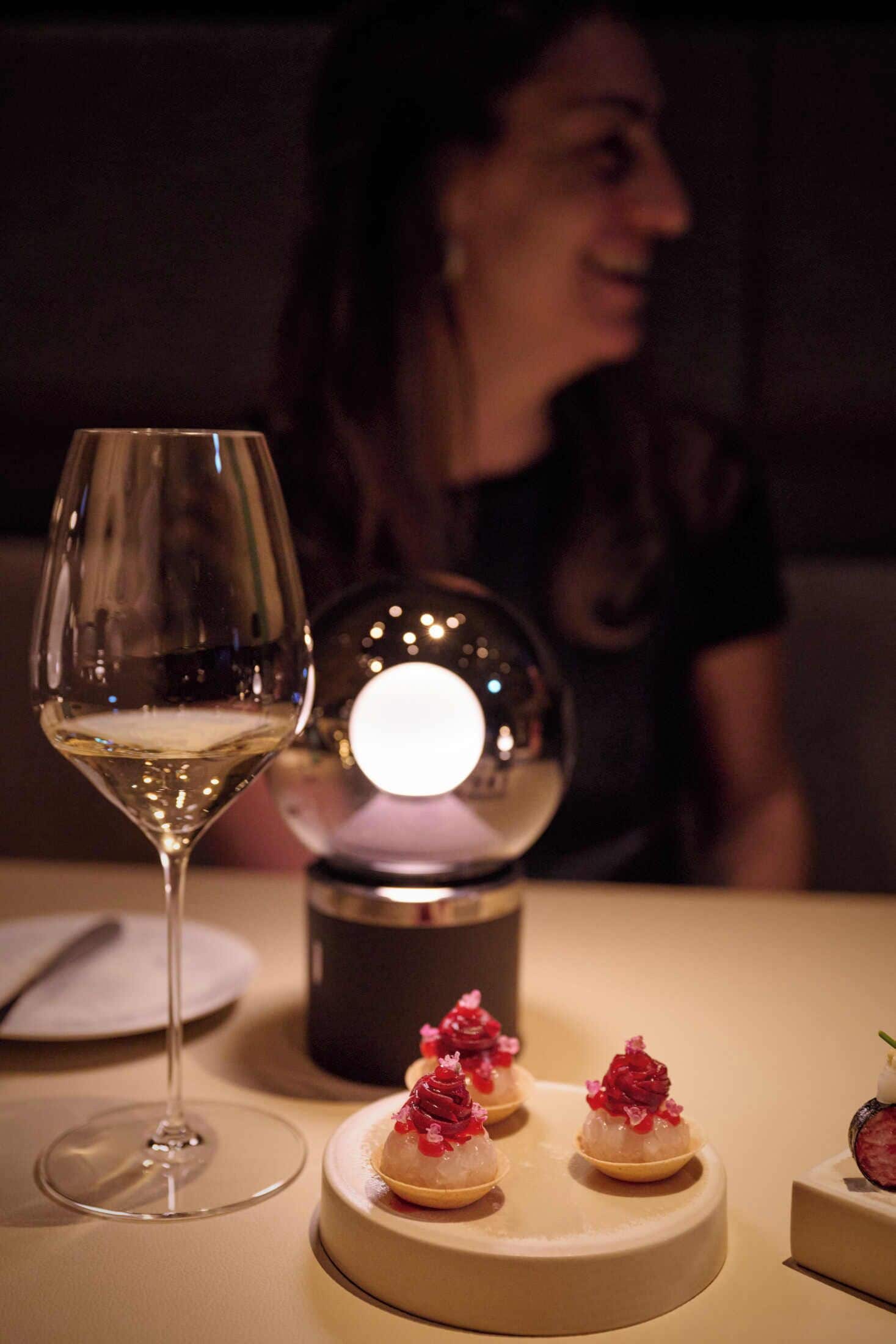 Ein Tisch mit einem Weinglas und drei kleinen Desserts auf einem runden Teller, im Hintergrund eine Kerze.
