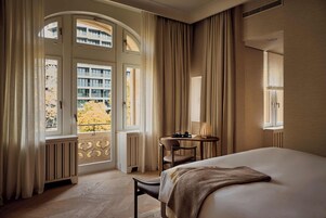 Une chambre d’hôtel avec un grand lit, une petite table avec une chaise, et une vue sur un immeuble par la fenêtre.