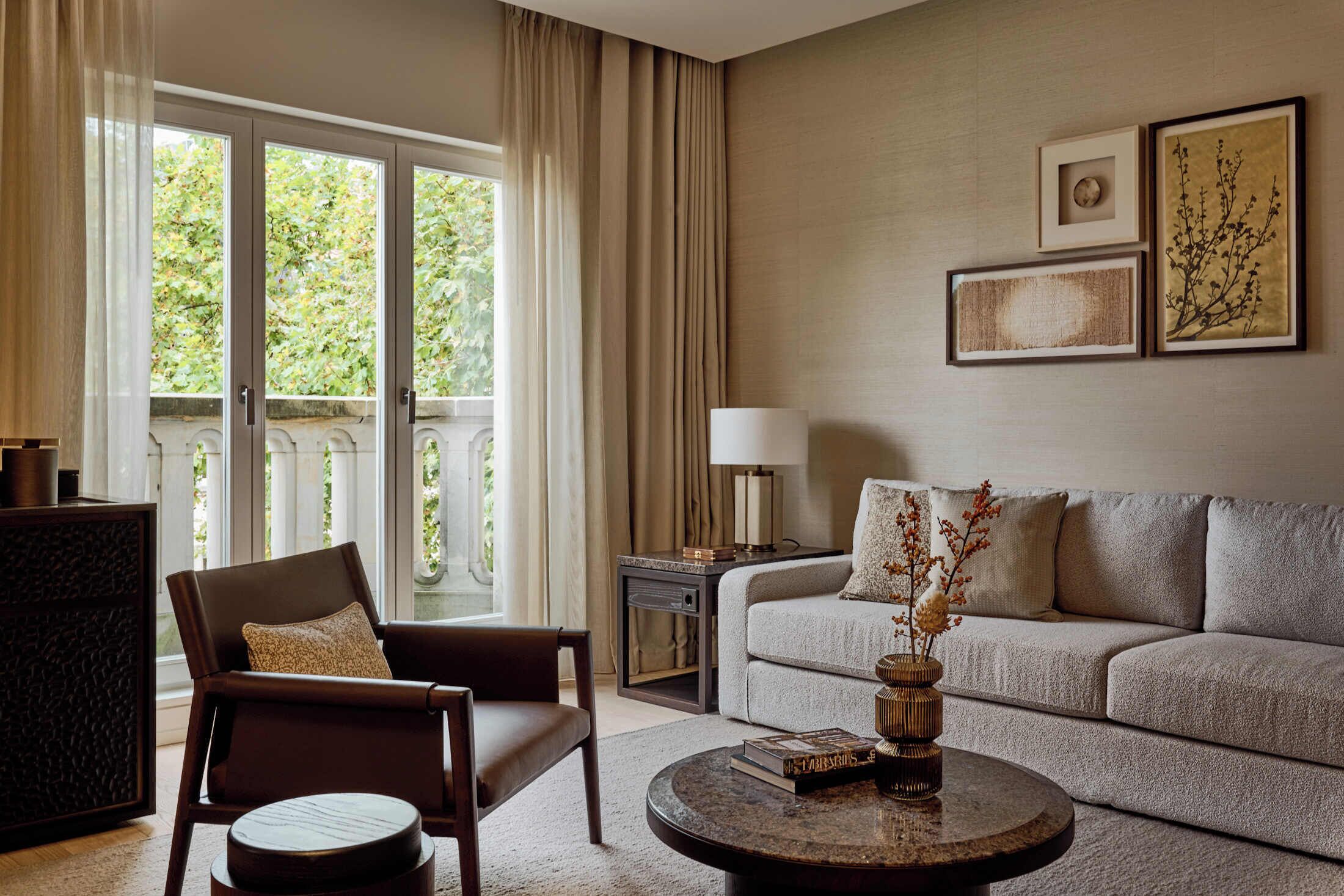 Una sala de estar moderna con un sofá, un sillón y una mesa de centro. Hay una ventana con cortinas, una lámpara y obras de arte enmarcadas en la pared.