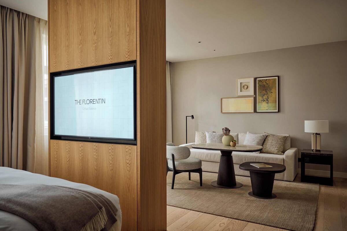 A hotel room with a bed, a TV displaying "THE FLORETIN Urban Retreat", a sofa, a small table, and a nightstand with a lamp.